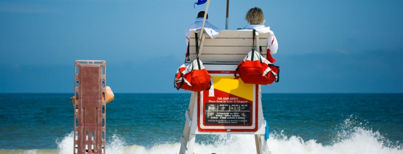 Two young men beach lifeguards watch people swimming in the blue ocean with waves crashing on a sandy beach.